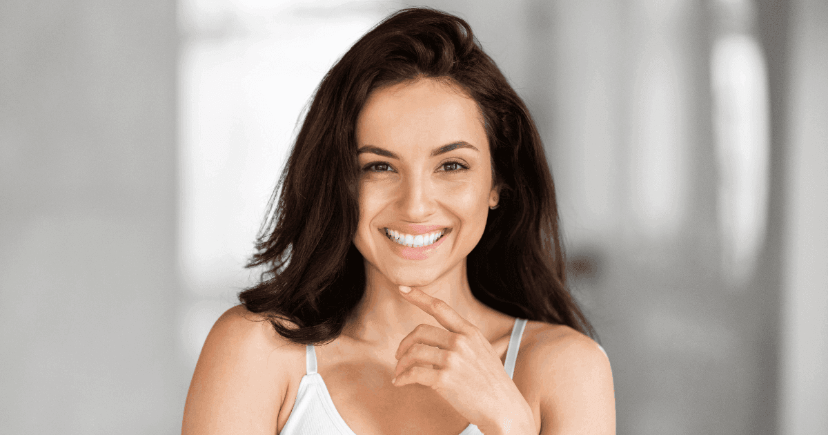 Smiling woman with long dark hair, touching her chin, against a light background.