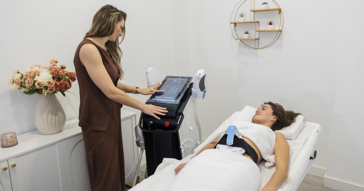 A woman operates a cosmetic treatment machine while another woman lies on a spa bed with a device strapped to her abdomen.